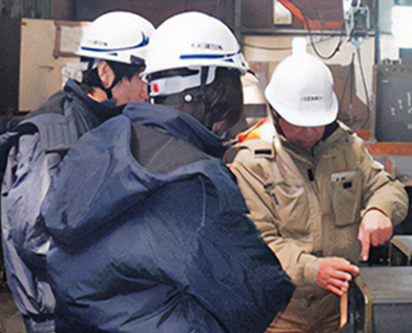 作業服を着てヘルメットを被った3人の工員が、作業現場で何かを確認している場面